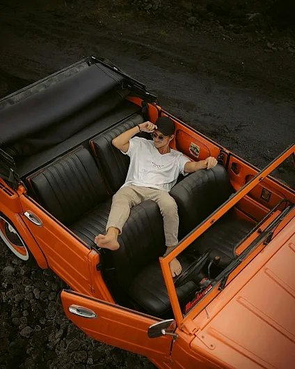 Eine Person entspannt sich barfuß in einem orangefarbenen Cabrio VW 181 Kübelwagen und liegt dabei quer über den schwarzen Ledersitzen. Die Szene vermittelt ein Gefühl von Muße und Abenteuer.