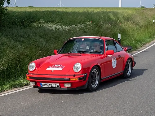 Ein leuchtend roter Porsche 911 im Rallye-Stil aus früheren Zeiten fährt an einem sonnigen Tag eine Landstraße entlang, hinter ihm erstrecken sich grüne Felder und Windräder.