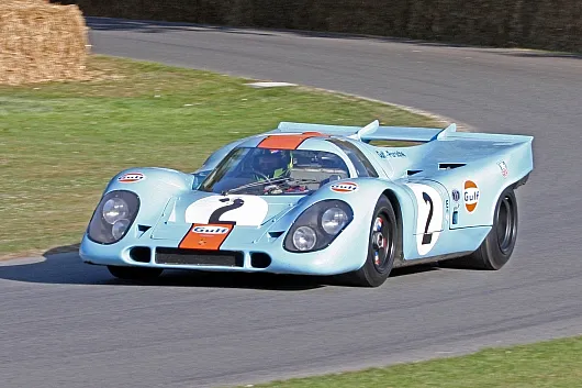 Ein hellblauer Oldtimer-Rennwagen Porsche 917k mit einem Gulf-Logo, der Startnummer 2 und eleganten Kurven rast über eine Rennstrecke, die von grünem Gras und einer Kurve aus Heuballen umgeben ist.