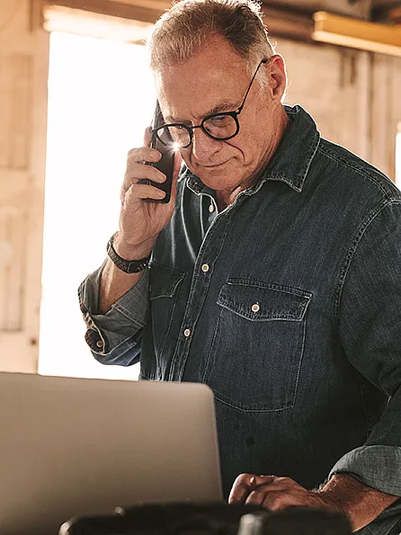 Ein Mann in einem Jeanshemd und mit Brille steht in einer Werkstatt, telefoniert und schaut dabei auf einen Laptop.