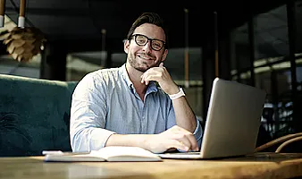 Ein junger Mann mit Brille und in einem blauen Hemd sitzt vor seinem Laptop und lächelt in die Kamera