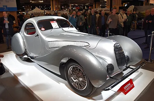 Ein eleganter silberner Oldtimer, der auf einer Plattform auf einer Automobilausstellung ausgestellt ist und von einer Menschenmenge umringt wird. Die Rundungen und Details des Wagens strahlen Eleganz und Nostalgie aus.