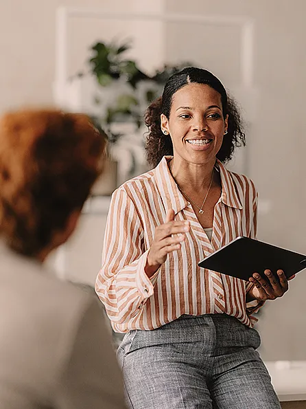 Eine Frau mit einem Tablet unterhält sich mit zwei sitzenden Personen. Sie lächelt, trägt ein gestreiftes Hemd und Jeans, im Hintergrund ist eine Pflanze zu sehen, was eine freundliche und professionelle Atmosphäre vermittelt.