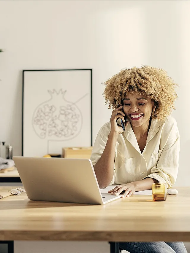 Person mit lockigem Haar telefoniert an einem Tisch mit Laptop und Getränk; im Hintergrund hängt ein Bild mit Granatapfelmotiv.