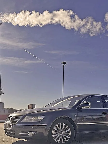 Eine schwarze VW Phaeton D1 3.0 TDI Individual 2007 parkt unter einem klaren blauen Himmel mit einer hellen Sonne in der Ecke. Im Hintergrund steigt Rauch aus hohen Industrieschornsteinen auf.