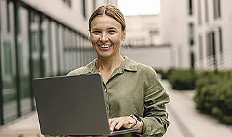Eine junge Frau mit zusammengebundenen Haaren und einer Bluse steht vor einem Bürogebäude und hält lächeln ihren Laptop im Arm
