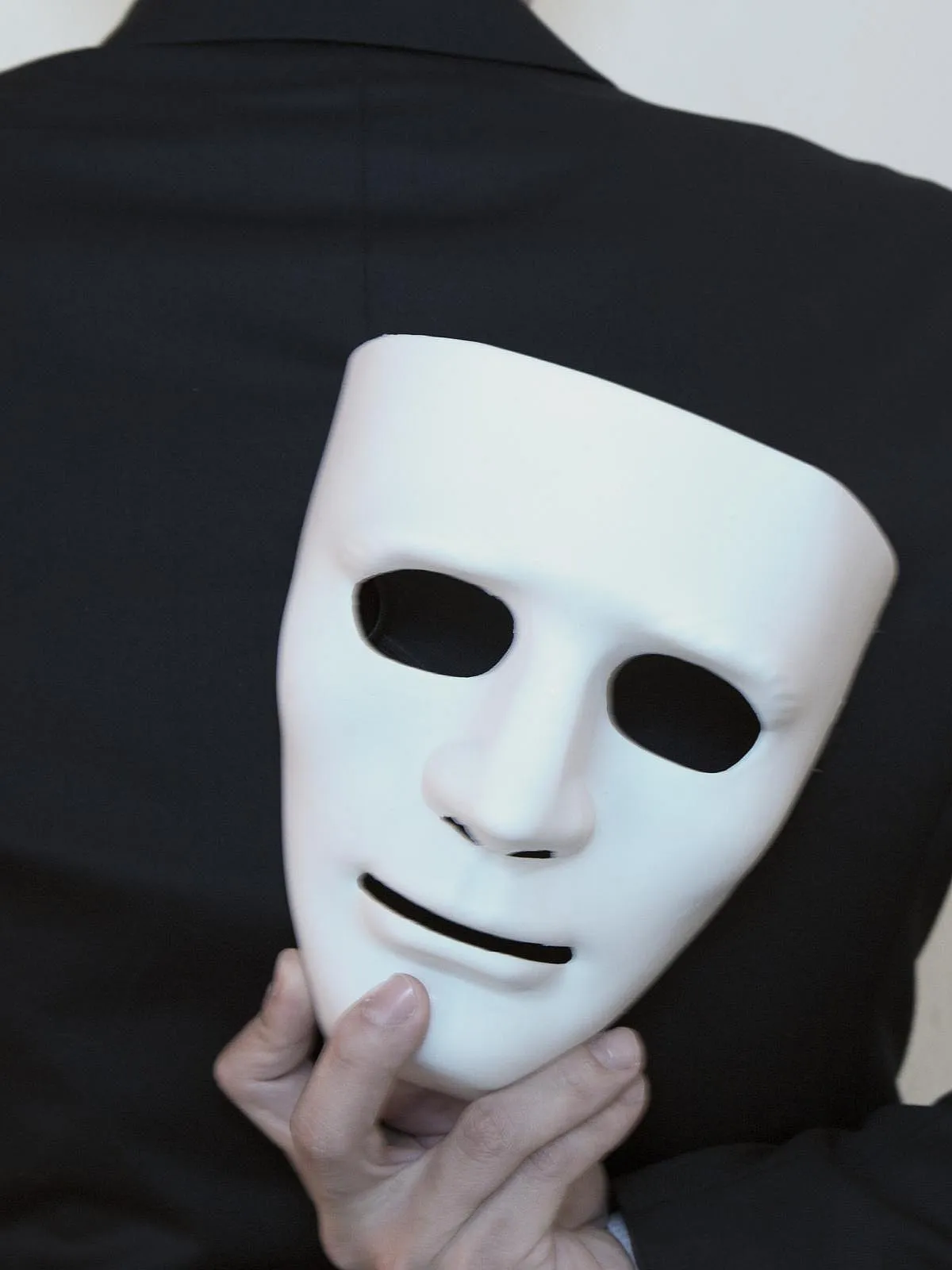 Eine Person in einem schwarzen Anzug hält eine weiße Maske hinter ihrem Rücken. Die Maske ist teilweise verdeckt und die Person ist von hinten zu sehen.