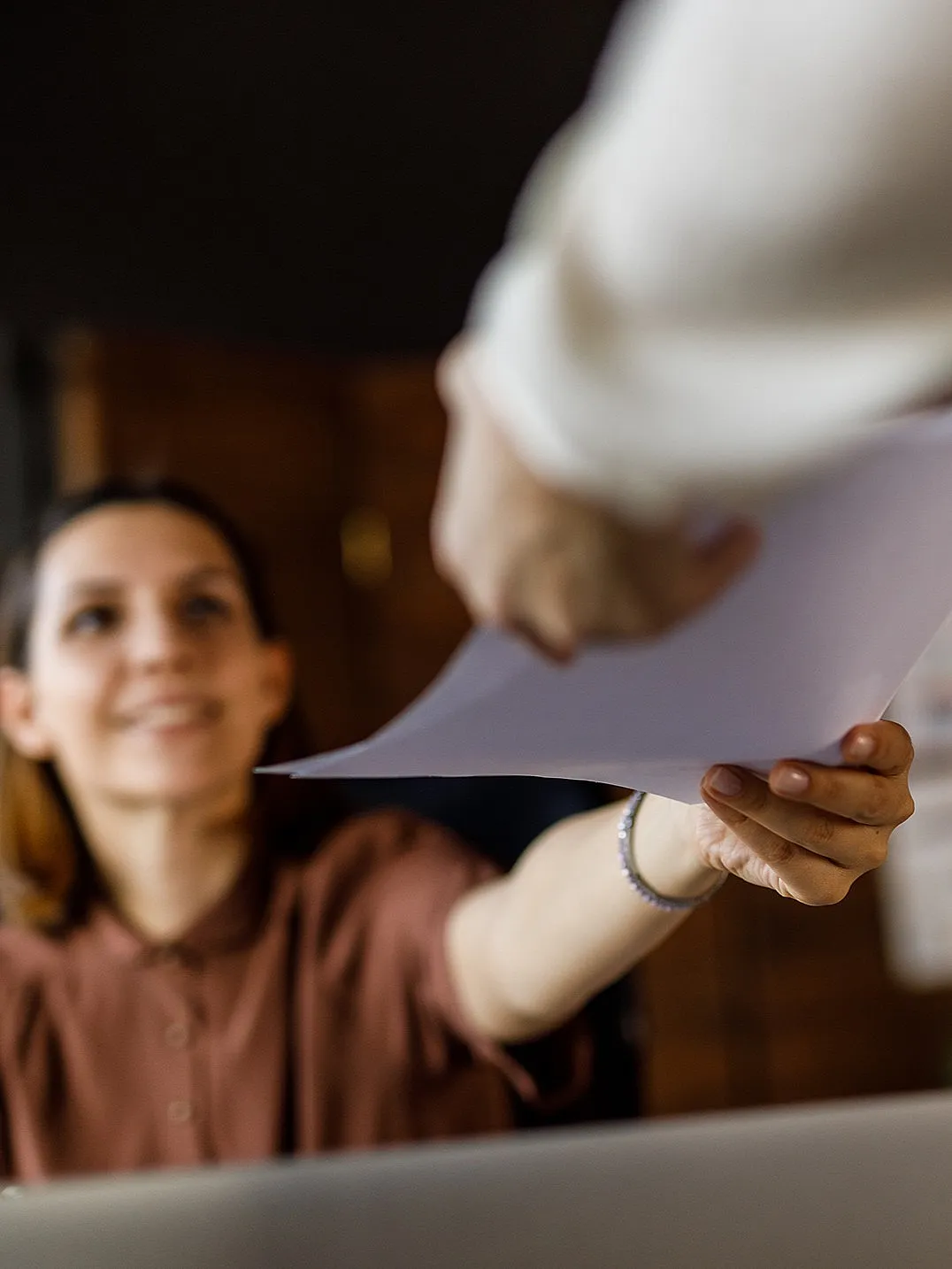 Eine lächelnde Frau, die an einem Schreibtisch sitzt, erhält Unterlagen von einer anderen Person. Büroumgebung mit Pflanzen und Dokumenten an einer Holzwand, die Teamarbeit vermittelt.