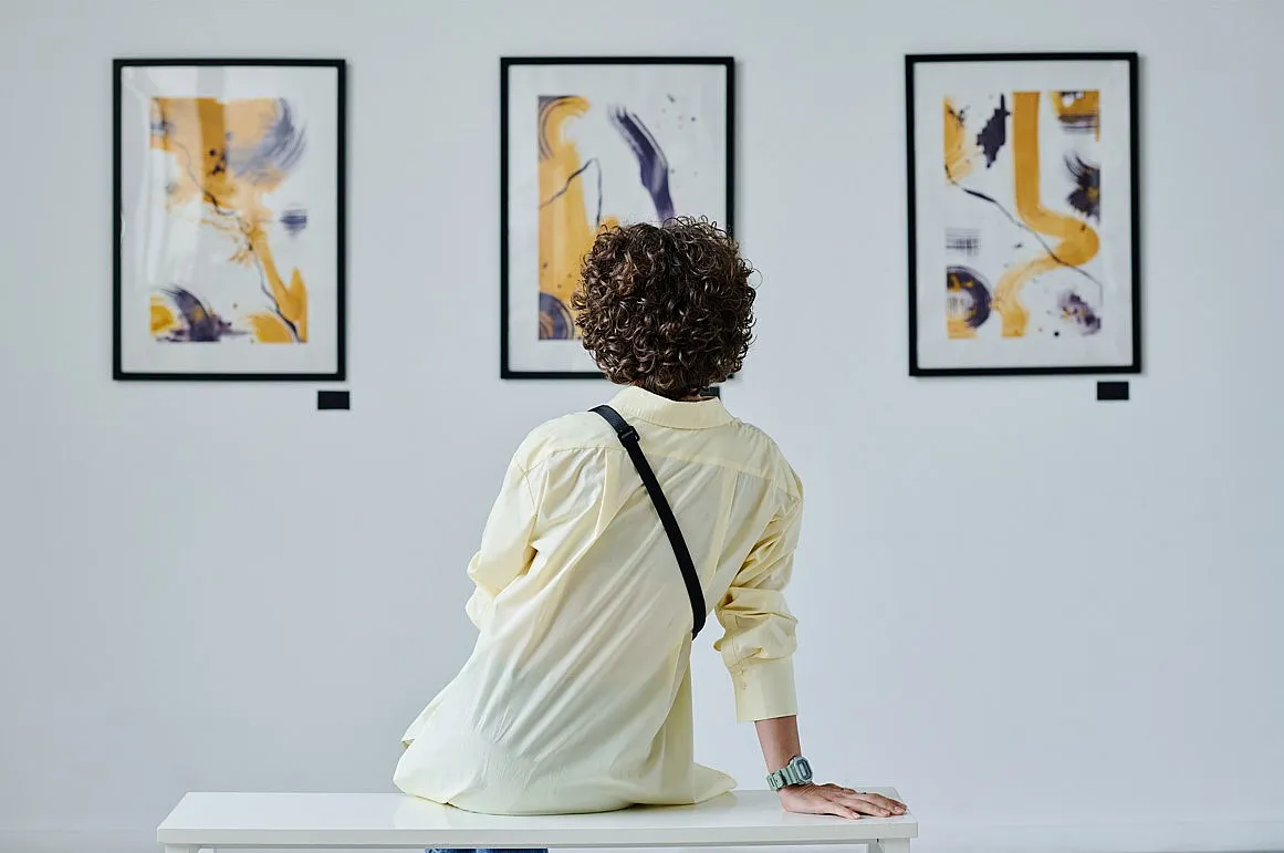 Eine Person sitzt auf einer Bank in einer Kunstgalerie und betrachtet drei abstrakte Gemälde an der Wand. Die Gemälde zeigen eine Mischung aus Gelb, Schwarz und Weiß mit verschiedenen Pinselstrichen und Formen. Die Person trägt ein helles Hemd und hat lockiges Haar