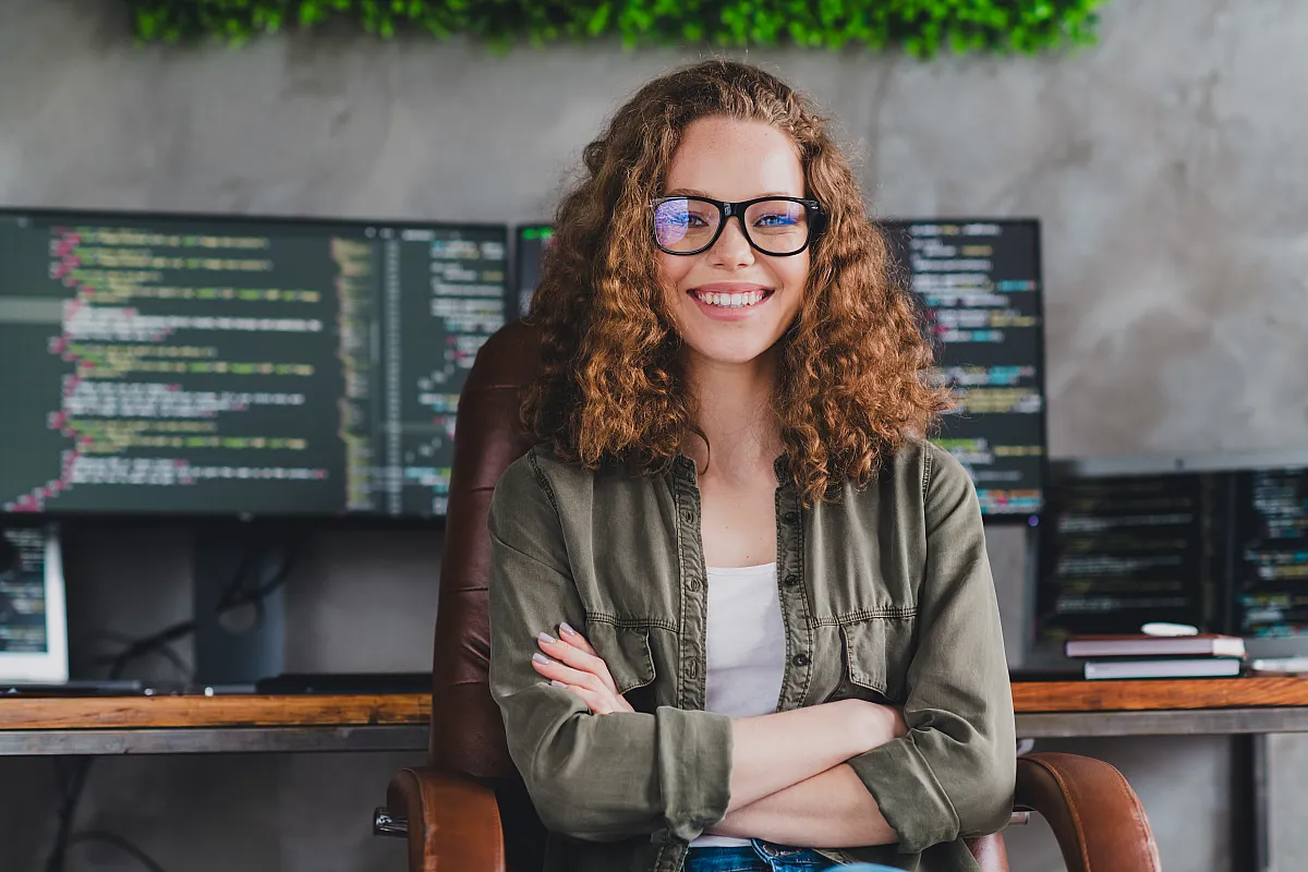 Eine lächelnde Person mit lockigem Haar und Brille sitzt selbstbewusst auf einem Bürostuhl. Hinter ihr zeigen Computerbildschirme eine Programmiersoftware. Hell und konzentriert.