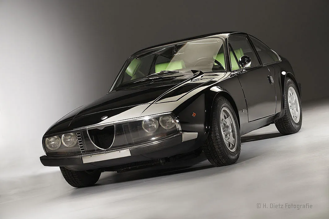 Ein schwarzer Alfa Romeo Montreal von 1970, fotografiert in einem Studio. Das Auto hat runde Scheinwerfer, einen verchromten Kühlergrill und glänzende Karosserie.