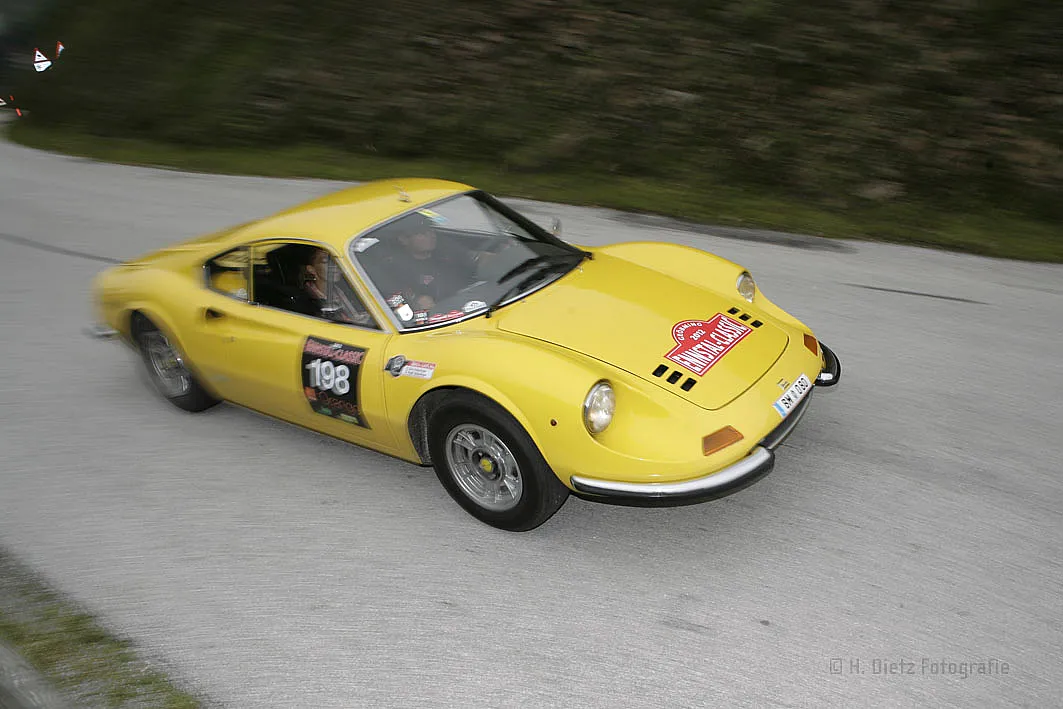Ein gelbes, klassisches Sportauto mit der Nummer 198, das an einem Rennen teilnimmt. Das Auto fährt auf einer asphaltierten Straße mit grüner Landschaft im Hintergrund.