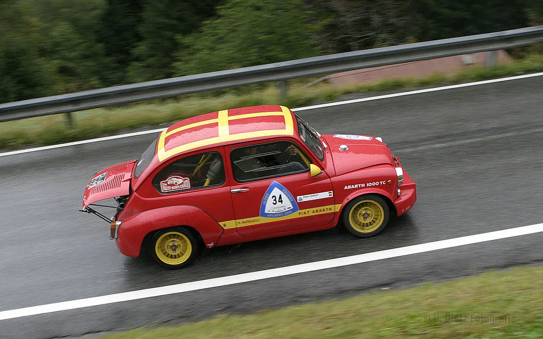 Ein rotes Rennfahrzeug, ein Fiat Abarth 1000 TC, fährt auf einer nassen Straße. Das Auto hat die Startnummer 34 und gelbe Felgen. Auf der Motorhaube und dem Dach sind gelbe Streifen zu sehen.