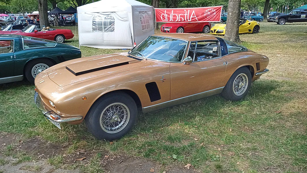 Ein klassisches Auto, ein Iso Grifo, geparkt auf einer Wiese bei einer Autoshow. Das Auto hat eine bronzene Farbe mit schwarzen Akzenten und Speichenrädern.