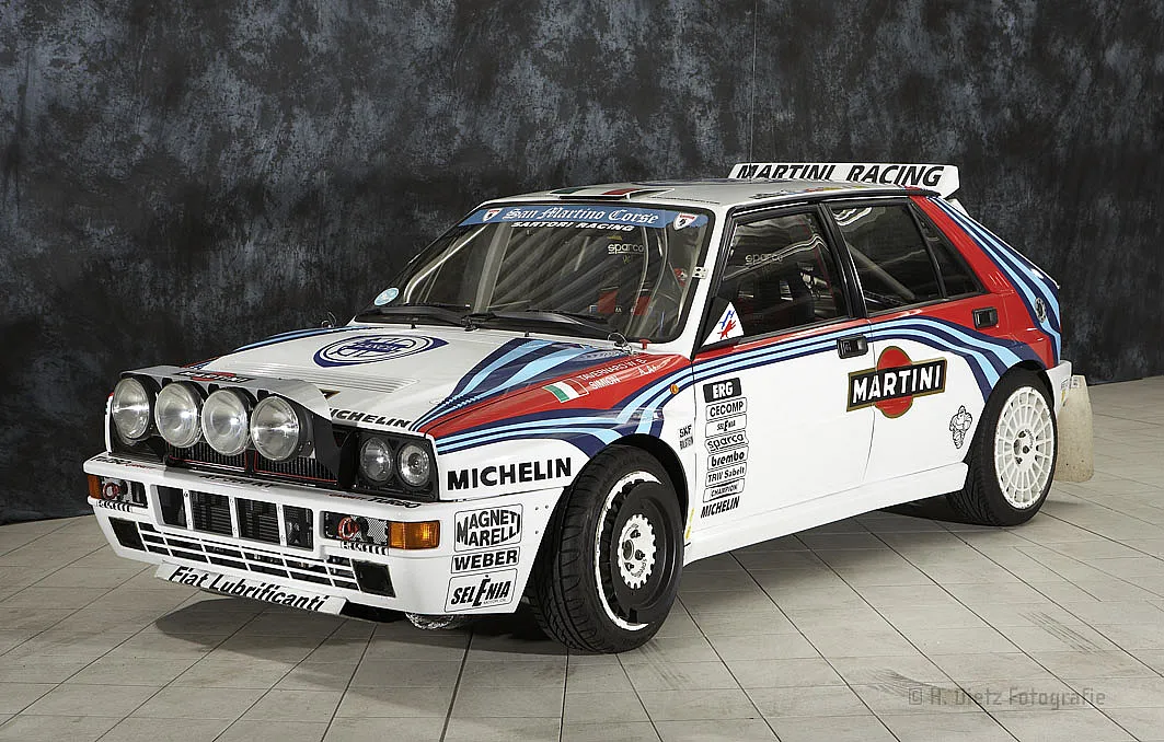 Ein weißer Rallye-Wagen mit blauen und roten Streifen, gesponsert von Martini Racing. Das Auto hat mehrere Aufkleber von Sponsoren wie Michelin und Magneti Marelli. Es ist auf einem grauen Fliesenboden in einem Innenraum geparkt.