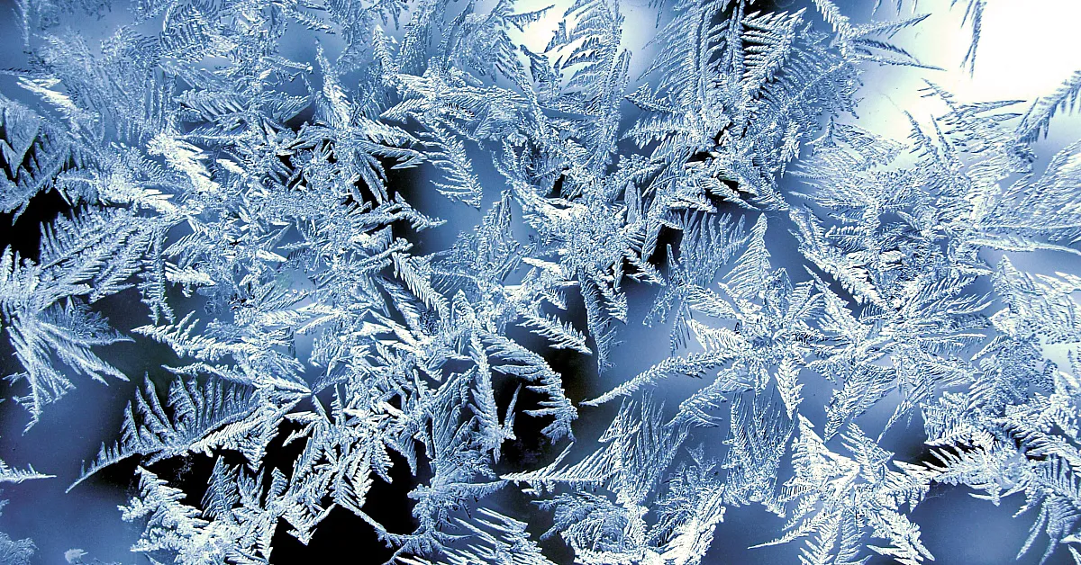 Ein Nahaufnahme von Frostkristallen auf einer Oberfläche. Die Frostkristalle haben komplizierte, federartige Muster und sind von Licht beleuchtet, was eine schöne und detaillierte Darstellung schafft.