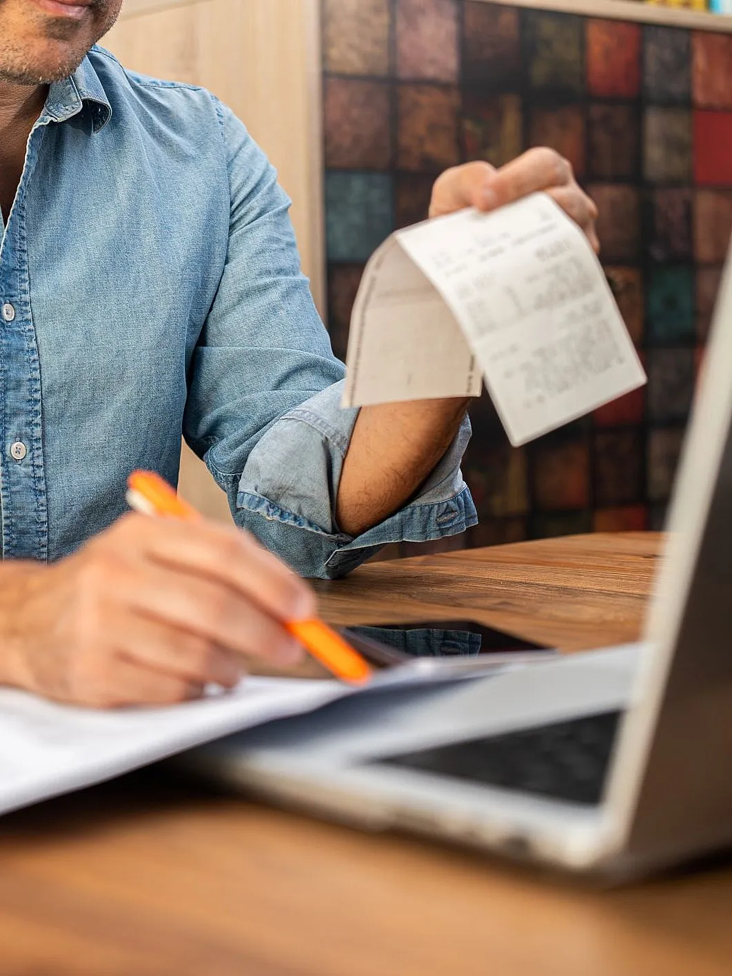 Ein Mann sitzt an einem Holztisch, trägt ein Jeanshemd und arbeitet am Laptop. In der einen Hand hält er einen Beleg oder eine Quittung, mit der anderen schreibt er etwas auf ein Blatt Papier. Auf dem Tisch liegen mehrere Dokumente.